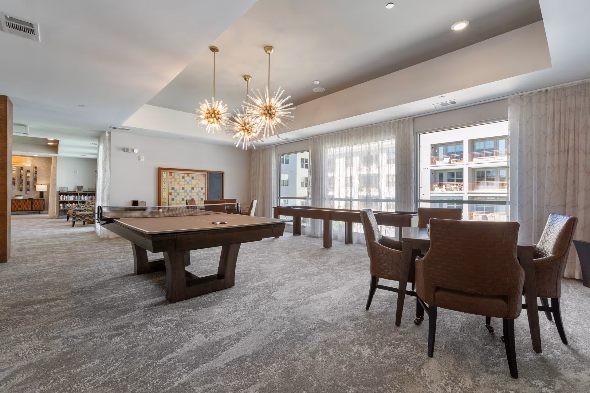 Bright communal game room with a pool table, shuffleboard table, and seating by large windows.