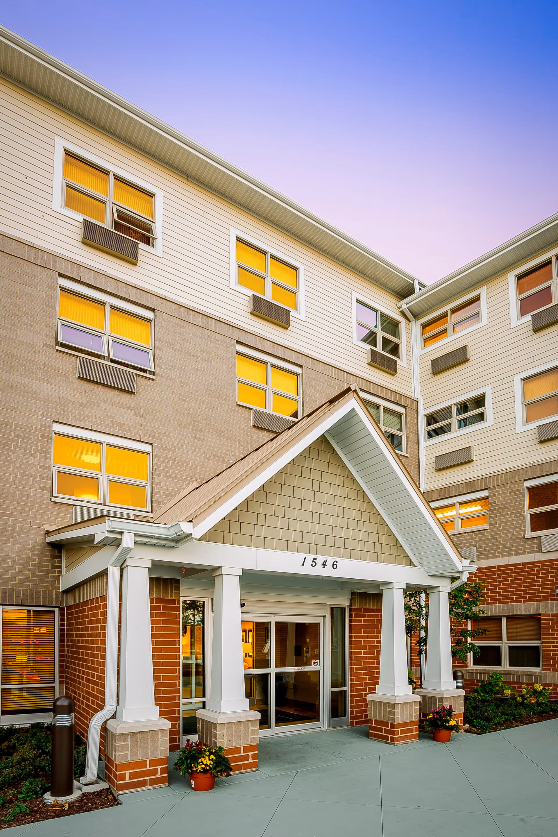 Entrance of a multi-story senior living facility building with a covered porch supported by white columns. The building has a combination of brick and siding exterior with multiple windows, some with air conditioning units. There are potted plants near the entrance and a clear sky at dusk.
