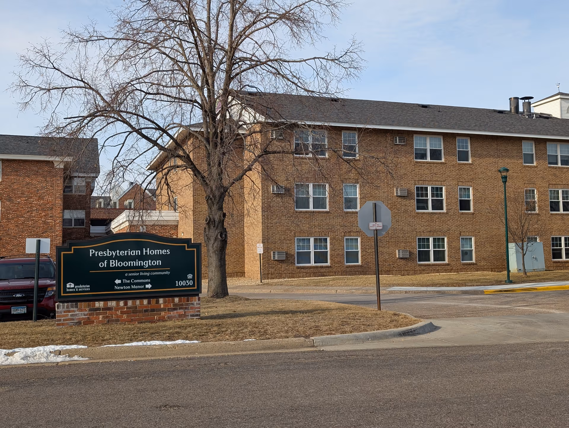 Exterior view of a brick senior living facility building with multiple windows. In front of the building is a large green sign reading 'Presbyterian Homes of Bloomington, a senior living community, The Commons, Newton Manor, 10030'. There is a leafless tree near the sign and a parked red vehicle partially visible on the left. The sky is clear with some light clouds.