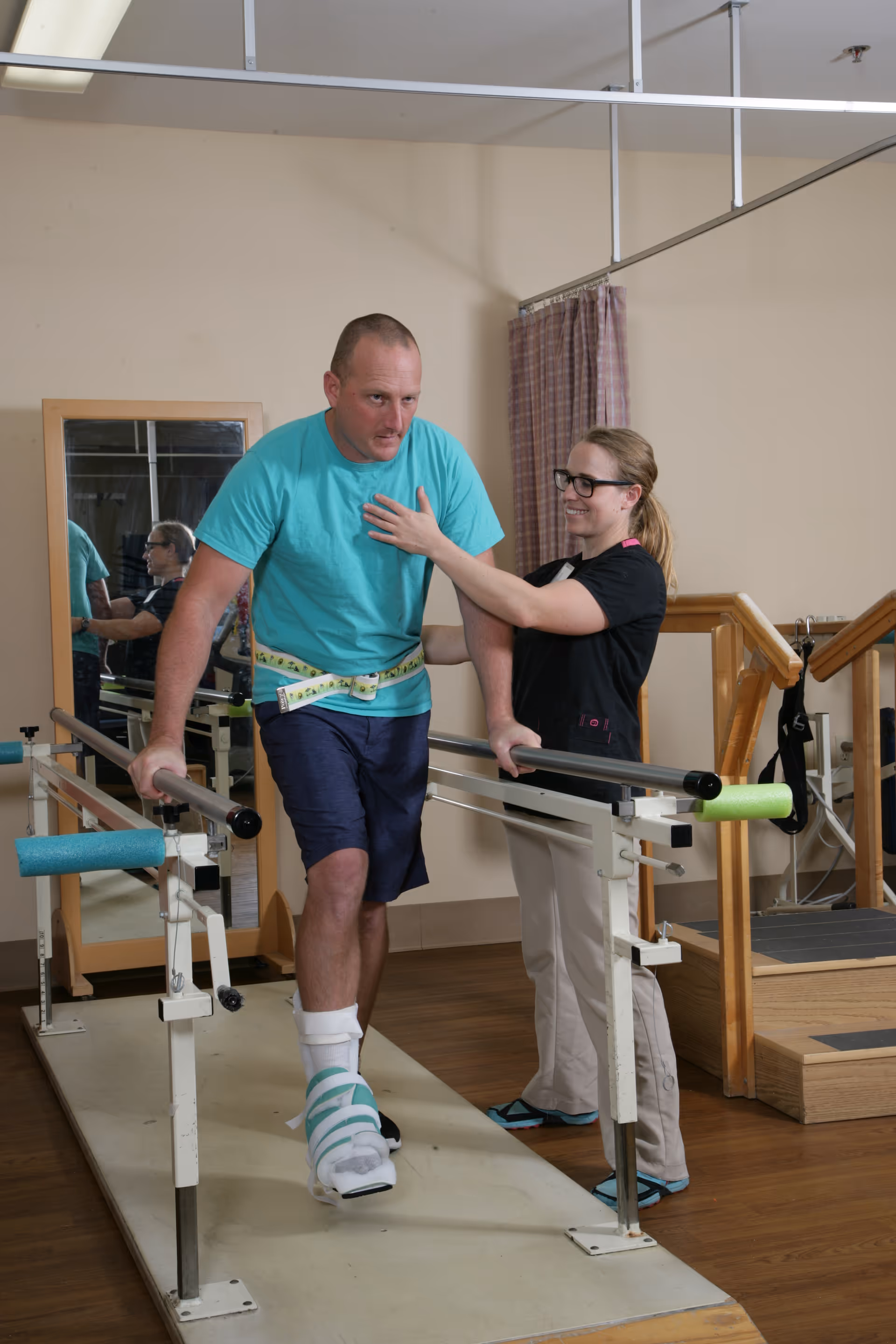 A man with a leg brace is walking between parallel bars for physical therapy, assisted by a female therapist who is supporting him and smiling. The setting appears to be a rehabilitation or therapy room with wooden flooring and exercise equipment.