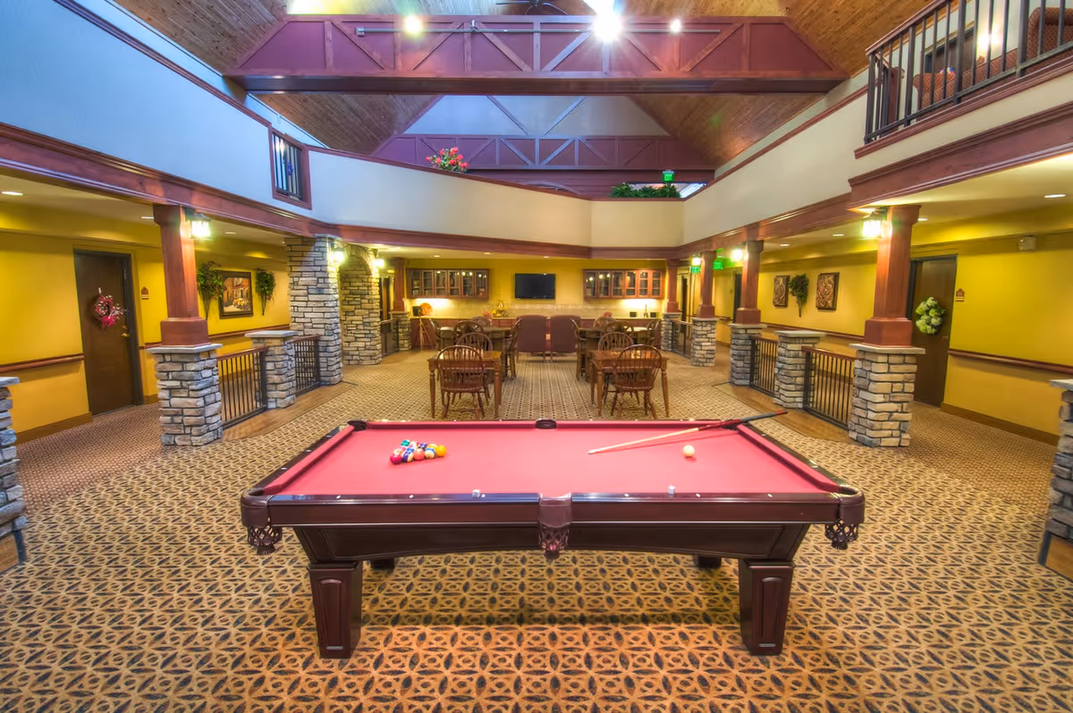 Interior view of a senior living facility common area with a red pool table in the foreground, several wooden tables and chairs in the background, stone pillars, yellow walls, and a high ceiling with exposed beams and lighting.