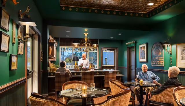 A cozy dining area with green walls and a decorative ceiling. There are round tables with wicker chairs, set with glasses and candles. Three elderly people are seated, two at a table on the right and one at the bar counter. A bartender stands behind the bar, serving drinks. The room is warmly lit with wall sconces and a chandelier above the bar.