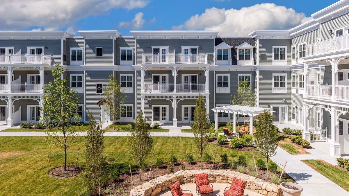 Courtyard with a circular stone seating area, lawn, young trees and pergola in front of a three-story senior living building with white balconies.