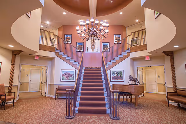 Interior view of a senior living facility lobby with a central carpeted staircase splitting into two upper flights. The area features a large chandelier, framed artwork on the walls, a grand piano with a horse sculpture on top, and seating areas with chairs along the walls. The space has warm lighting and a patterned carpet.