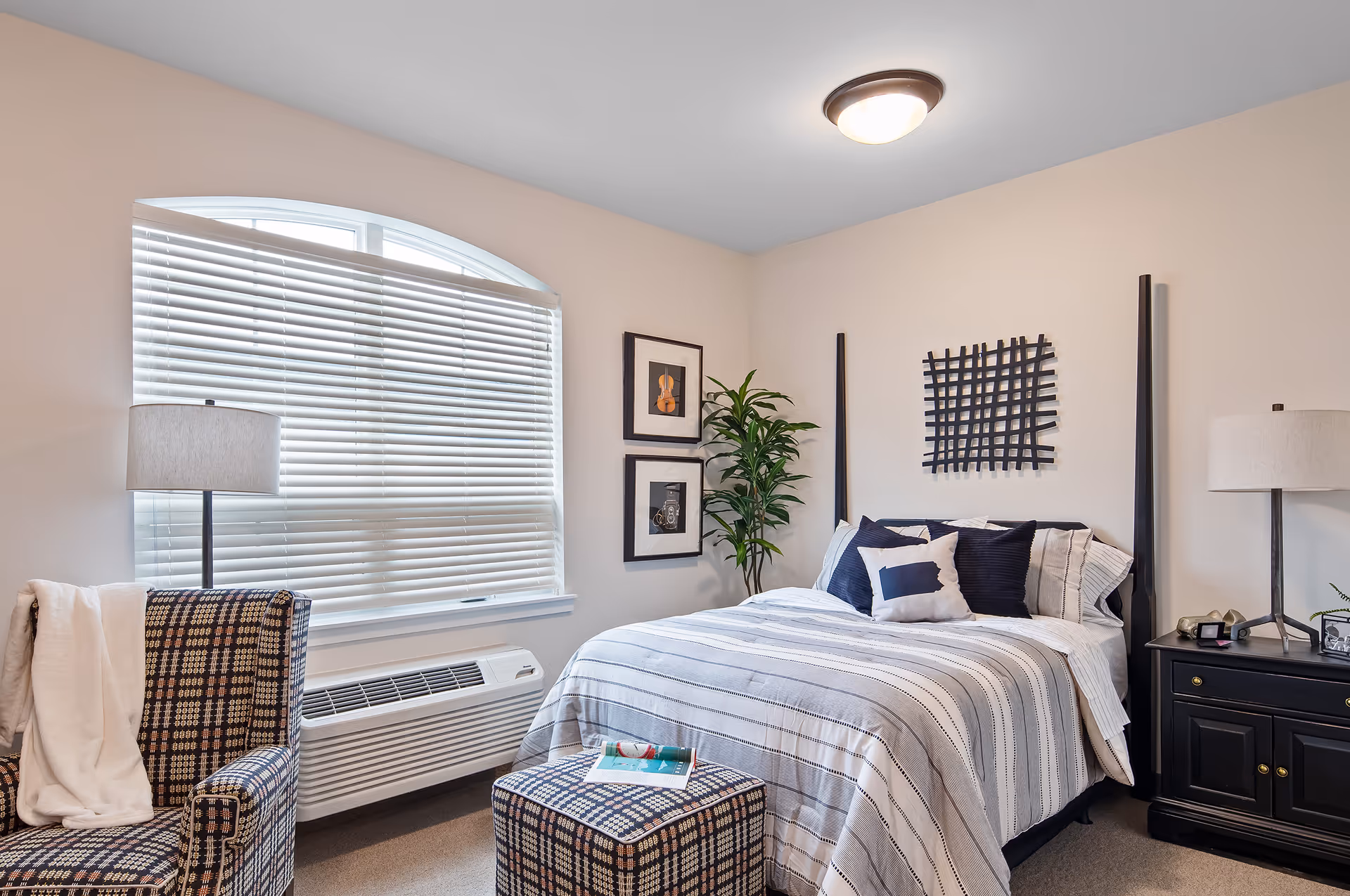 A neatly arranged bedroom with a bed dressed in striped bedding and multiple pillows. There is a black nightstand with a lamp on the right side of the bed. A patterned armchair with a white blanket draped over it and a matching ottoman with a magazine on top are positioned near a window with white blinds. Two framed pictures hang on the wall next to a tall green plant. The room has beige walls and a ceiling light fixture.