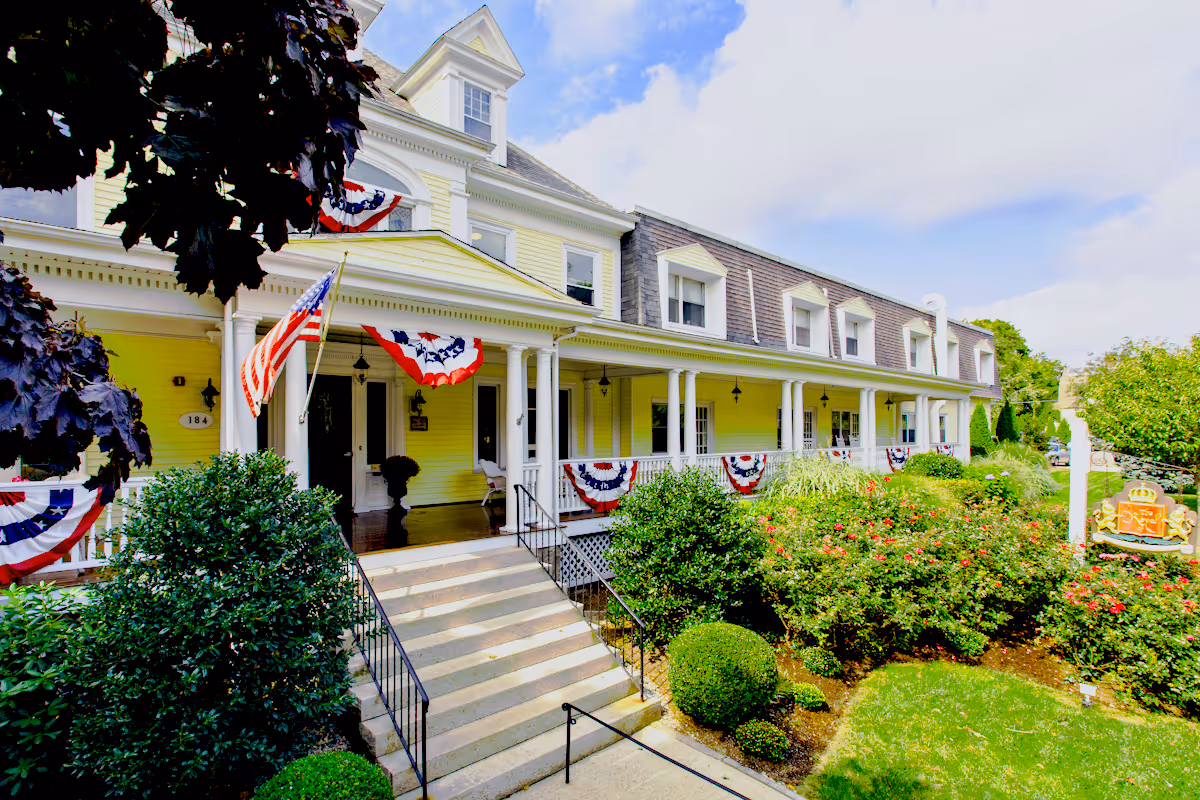 Front exterior view of a large yellow nursing center building with white columns and a porch decorated with American flag bunting. There are stairs leading up to the entrance, surrounded by well-maintained bushes and greenery under a partly cloudy sky.