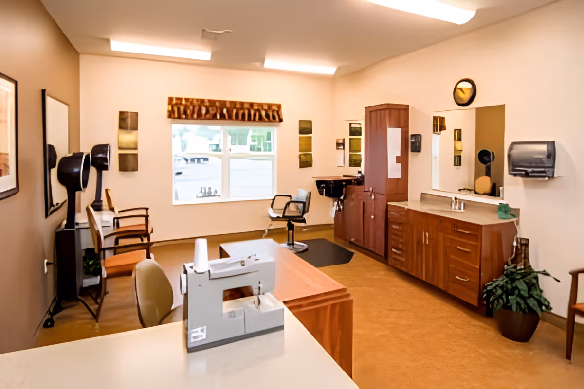 Interior view of a salon area in an assisted living facility with salon chairs, hair dryers, a sink, cabinets, a large mirror, and a window with a decorative valance. There is a sewing machine on a counter in the foreground and a potted plant near the cabinets.