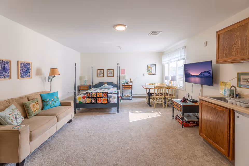 A bright and spacious studio apartment with a beige sofa adorned with colorful pillows on the left, a four-poster bed with a patchwork quilt in the center, a small dining table with four chairs near a window on the right, a wall-mounted flat-screen TV, and a kitchenette with wooden cabinets and a sink on the far right.