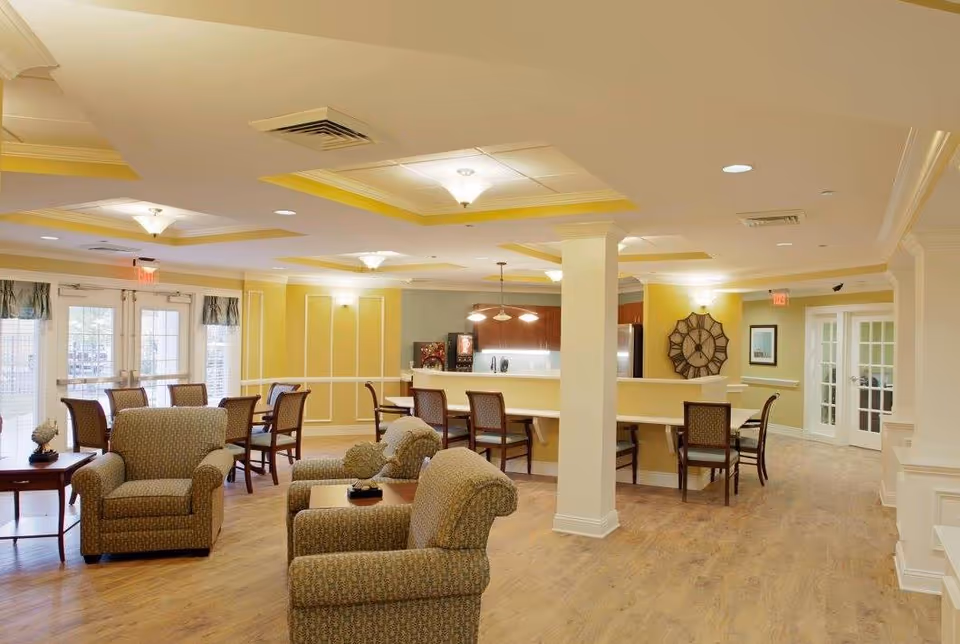 Spacious, well-lit common lounge with upholstered armchairs, small tables, dining chairs and a service counter.