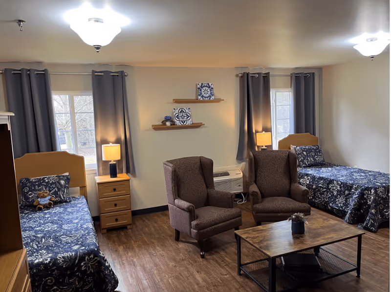 A cozy assisted living bedroom with two single beds, each with blue floral bedding and a pillow. Between the beds is a wooden nightstand with a lamp. Two brown upholstered armchairs face a wooden coffee table with a small plant on it. The room has two windows with gray curtains and two decorative shelves on the wall with blue and white decor items. The floor is wood, and the walls are light-colored.