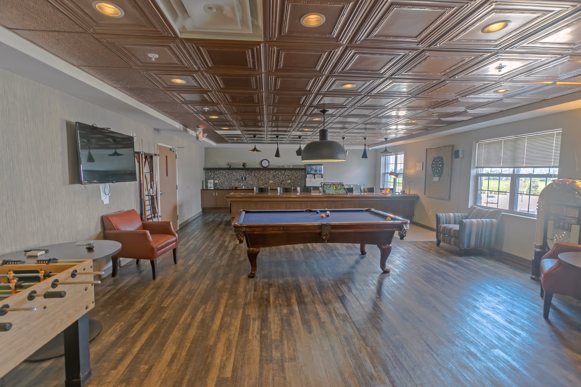 A recreational room with a pool table in the center, a foosball table on the left, and several chairs around the room. There is a large flat-screen TV mounted on the left wall, a dartboard on the right wall, and a counter with bar stools at the back. The room has wooden flooring and a decorative ceiling with recessed lighting.
