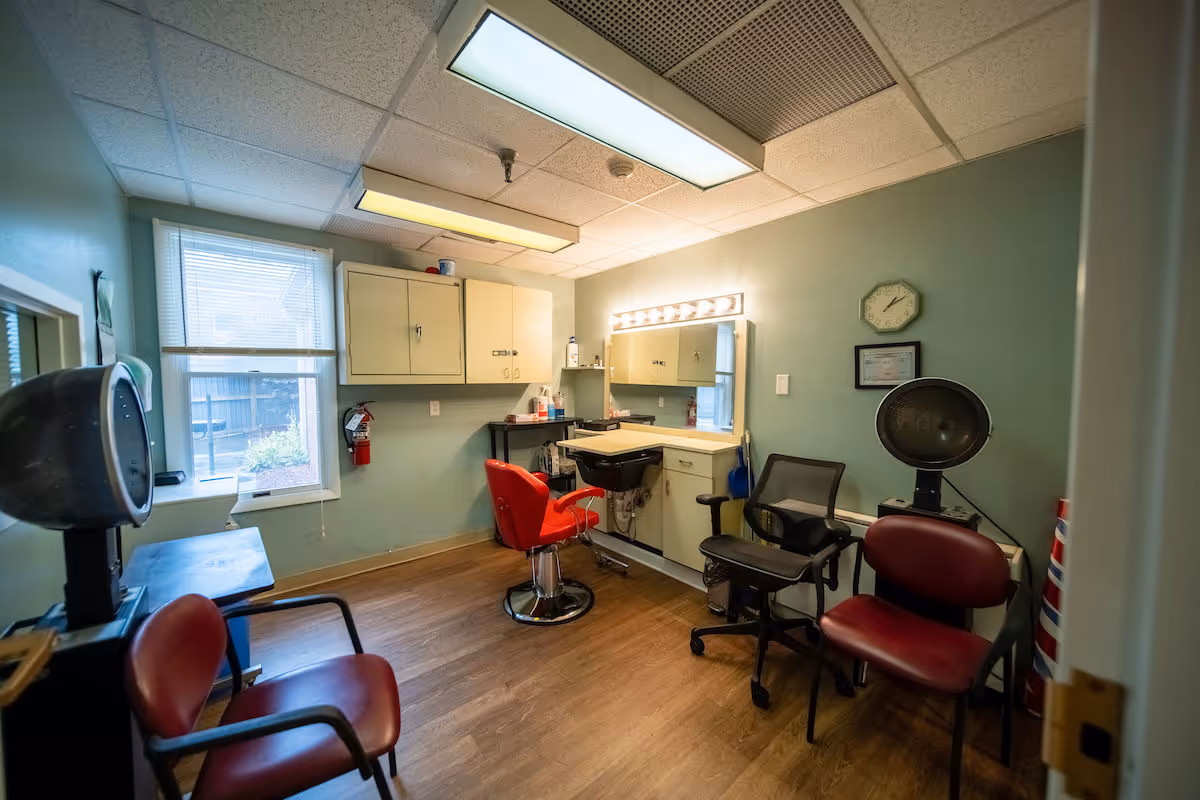 A small room with light green walls and wood flooring, furnished as a hair salon with a red salon chair in front of a mirror and sink, additional chairs for waiting, a hair dryer, and cabinets on the wall. A window with blinds lets in natural light.