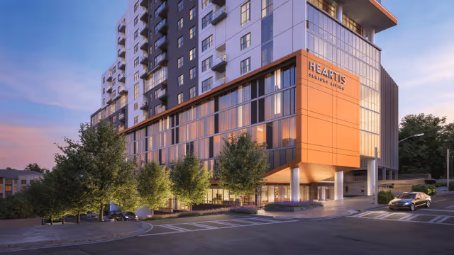 Exterior view of a modern multi-story senior living facility named Heartis Buckhead Senior Living during sunset, featuring large windows, an orange accent panel with the facility name, surrounding trees, and a car parked near the entrance.