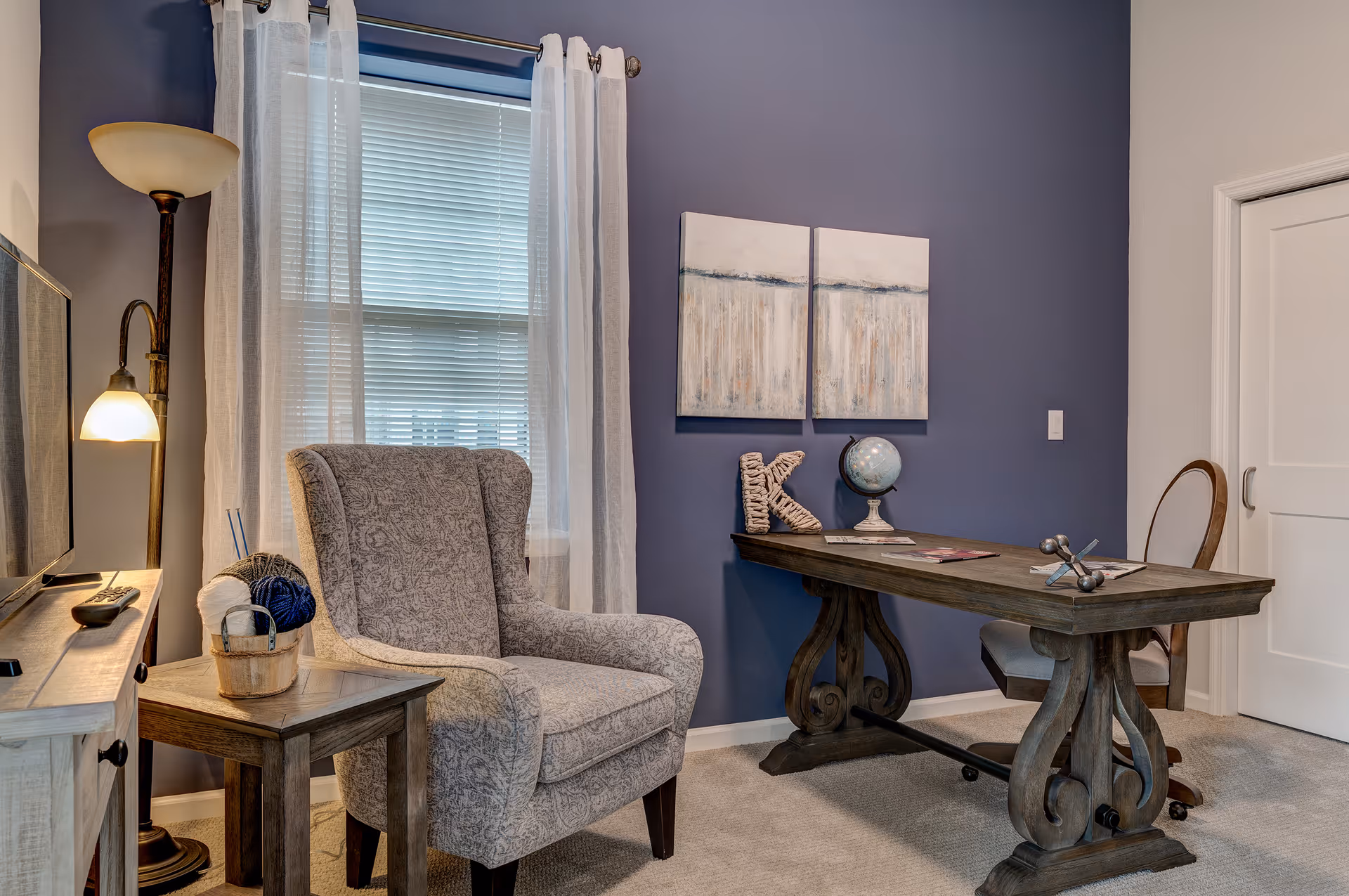 A cozy interior room with a patterned armchair next to a small wooden side table holding a basket with yarn balls. A tall floor lamp with two lights stands behind the chair. A wooden desk with decorative items including a globe, a letter K, and magazines is positioned against a blue accent wall with two abstract paintings. A cushioned chair is placed at the desk, and a window with white curtains and blinds is visible behind the armchair.