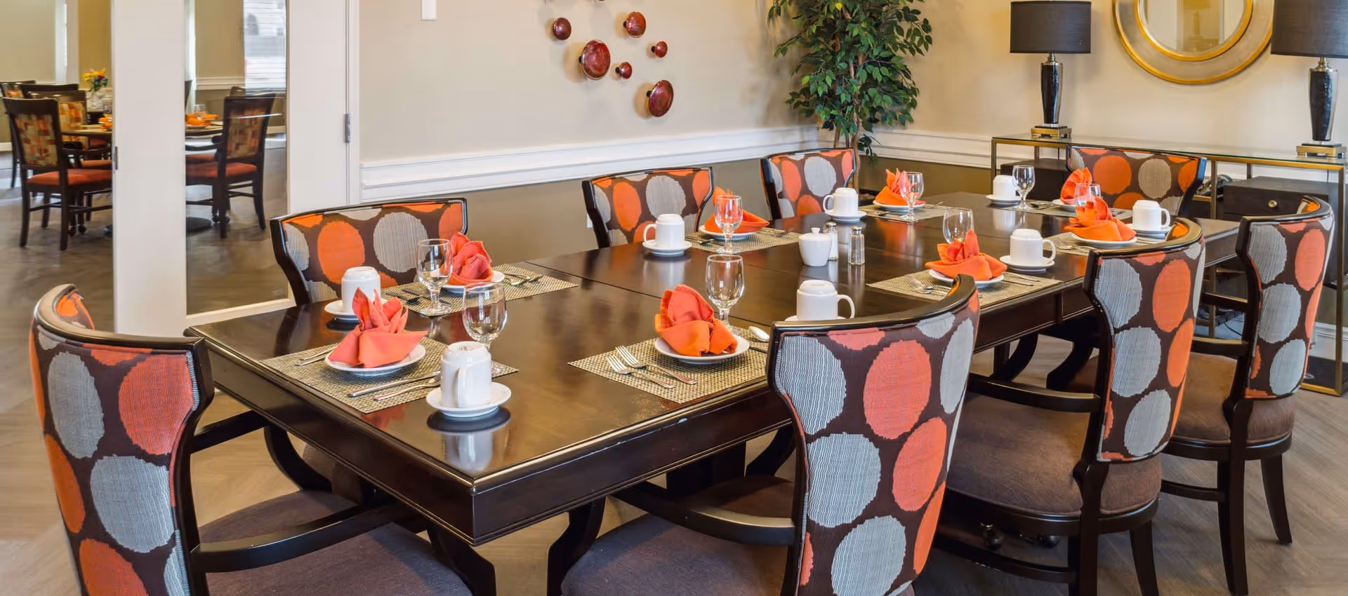 A dining room with a dark wooden table set for eight people. Each place setting includes a white cup and saucer, a wine glass, silverware, and an orange cloth napkin folded on a white plate. The chairs have a colorful upholstery with large orange and gray circles. The room features a decorative wall with red circular wall art, a green potted plant, two black table lamps with gold bases, and a round gold-framed mirror.