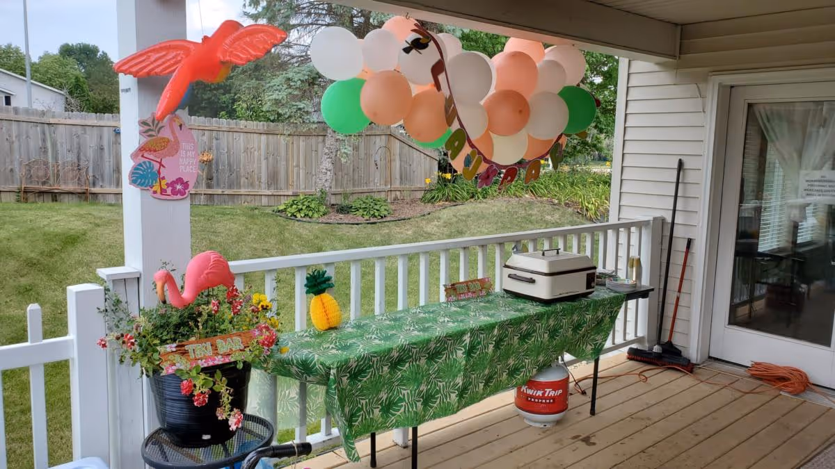 A covered outdoor patio area decorated with tropical-themed items including a pink flamingo, a pineapple decoration, and a green tablecloth with leaf patterns on a table. The table holds a small appliance and some plates. Above the table, there is a cluster of balloons in white, peach, and green colors with a banner. The patio overlooks a grassy yard with a wooden fence and some plants. Cleaning tools and an orange extension cord are visible near a glass door.