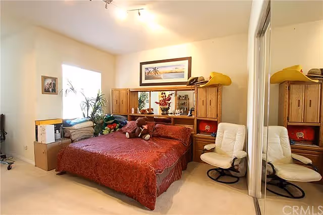 A bedroom with a large bed covered in a red patterned bedspread and several stuffed animals on it. Behind the bed is a wooden headboard with cabinets and shelves, decorated with a framed picture of a beach scene and various items. To the right of the bed is a white cushioned swivel chair, and a mirrored closet door reflects part of the room. A window with blinds and some plants are visible on the left side of the room.