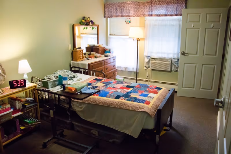A small bedroom with a single bed covered by a colorful patchwork quilt. There is a bedside table with a lamp and a digital clock showing 5:39. A wooden dresser with a mirror and various personal items is against the wall. A floor lamp stands near a window with white curtains and an air conditioning unit. The door to the room is open.
