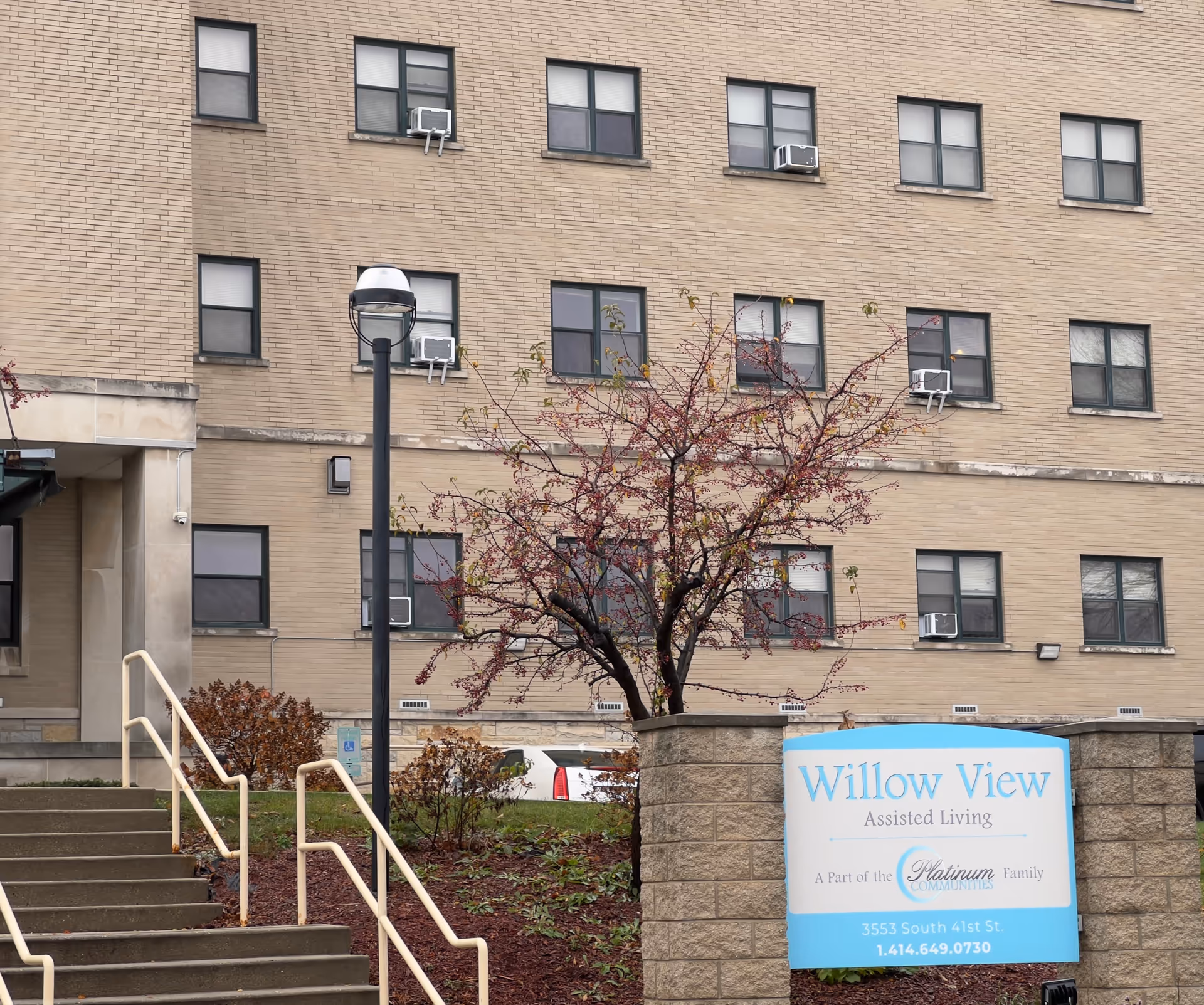Exterior view of a multi-story brick building with several windows, some with air conditioning units. In front of the building is a small landscaped area with a tree and bushes, a set of stairs with handrails leading up to an entrance, and a sign that reads 'Willow View Assisted Living, A Part of the Platinum Family Communities, 3553 South 41st St., 1.414.649.0730'.