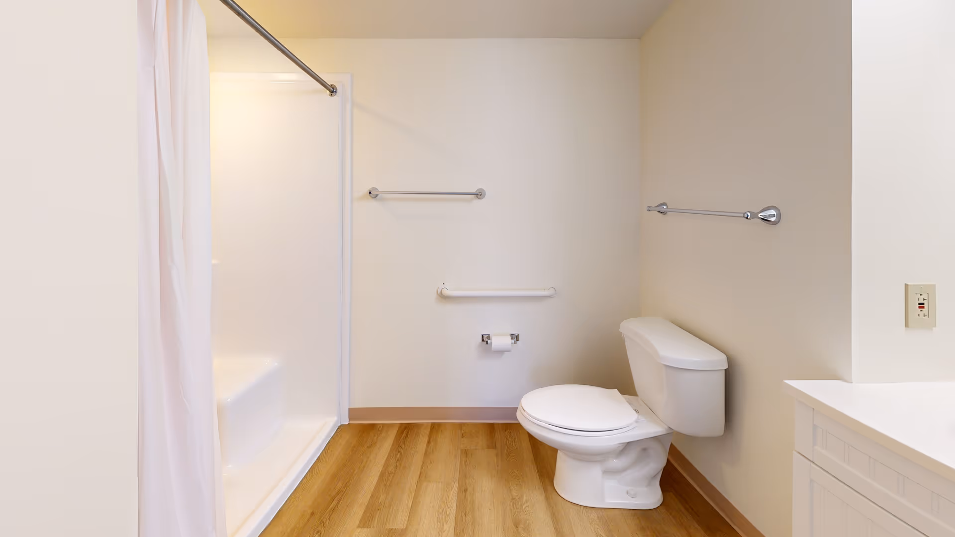 A clean bathroom with a white toilet, a shower with a white curtain, two towel bars on the walls, a grab bar near the toilet, and a wooden floor.