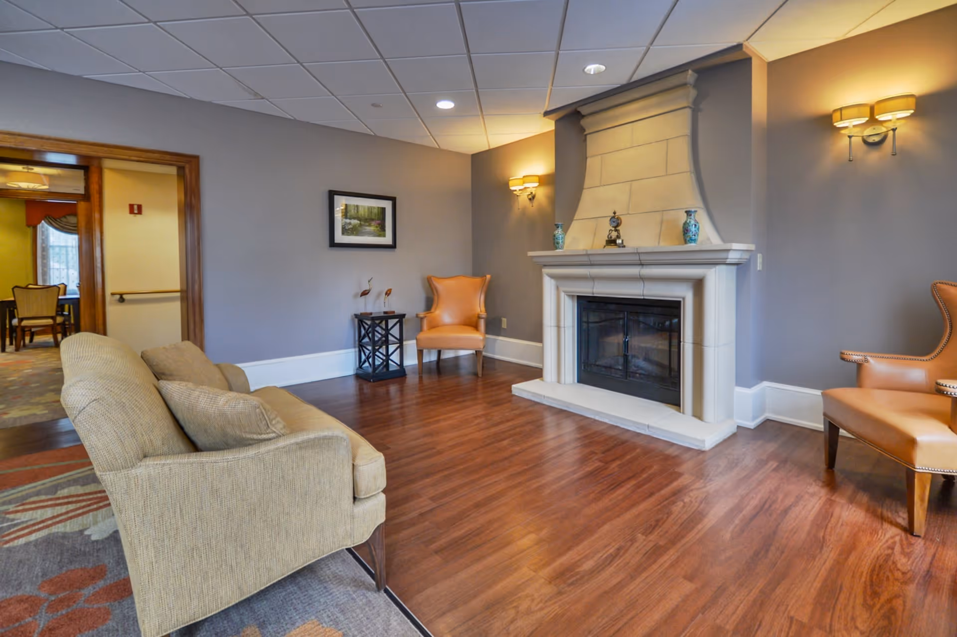A cozy living room area with a beige sofa and two orange armchairs arranged around a large stone fireplace. The room has wooden flooring, soft lighting from wall sconces, and a framed picture on the gray wall. In the background, there is a view into another room with dining chairs and a window.