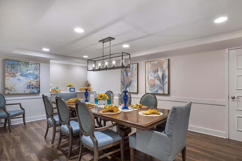 A well-lit dining room with a wooden table set for six people. The table has blue and white decorative items, yellow napkins, and a centerpiece with sunflowers. The chairs around the table have light blue upholstery. The room has wooden flooring, white walls, and three framed floral paintings on the walls. A modern rectangular chandelier hangs above the table.