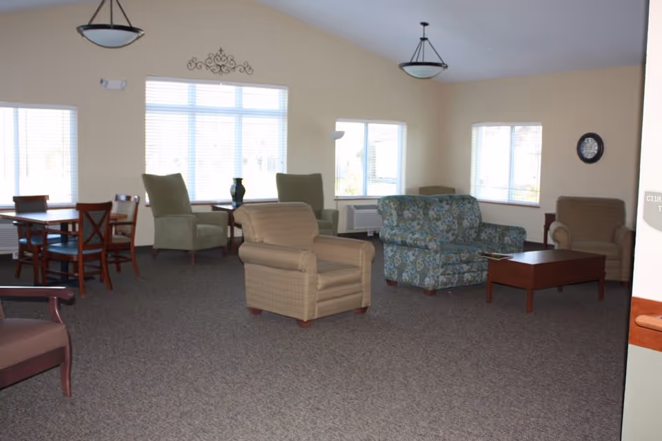 A spacious common area with multiple seating options including armchairs, a loveseat with floral upholstery, and a wooden coffee table. There is a dining table with chairs near the windows, which allow natural light to fill the room. The walls are light-colored, and the ceiling has two hanging light fixtures.