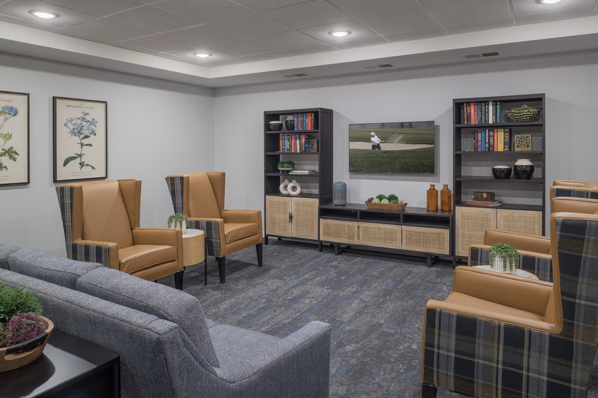 A cozy senior living common area with a gray sofa and four tan leather armchairs with plaid backs arranged around small round tables. Two black bookshelves and a matching TV stand with decorative items and books flank a wall-mounted flat-screen TV showing a golfer in a sand trap. The room has light gray walls, framed botanical prints, and a patterned carpet.