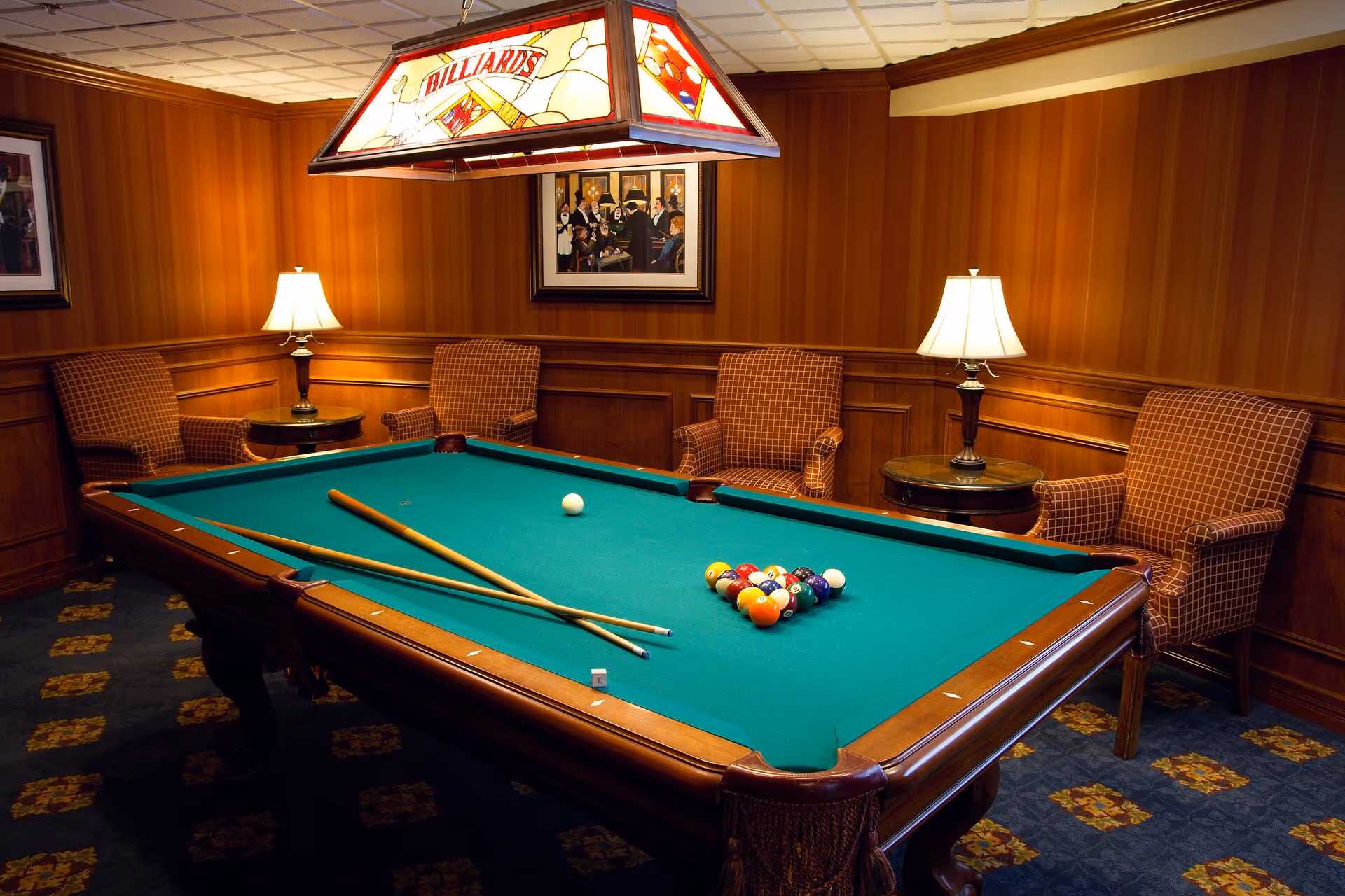 Wood-paneled game room featuring a billiards table with cues and racked balls, surrounded by armchairs and lamps.