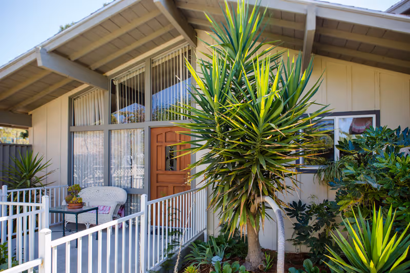 Front entrance of a single-story building with a wooden door, large windows, a small fenced porch with seating, and tropical plants.