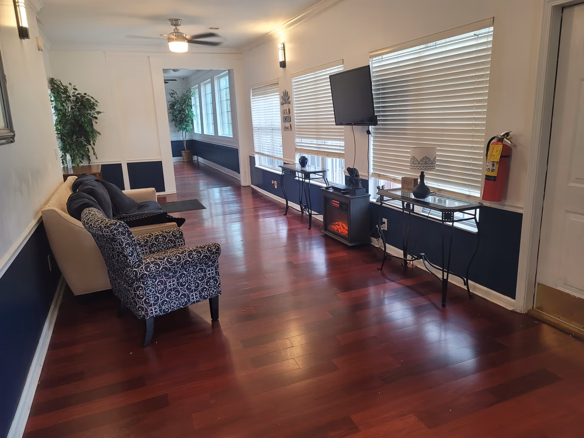 A long hallway with polished wooden floors and white walls with dark blue lower panels. On the left side, there is a beige sofa with dark cushions and a patterned armchair. On the right side, there are three windows with closed blinds, a wall-mounted TV, a small electric fireplace, two glass-top tables with decorative items, and a fire extinguisher mounted on the wall. The hallway is well-lit with wall sconces and ceiling lights, and there are potted plants at the far end.