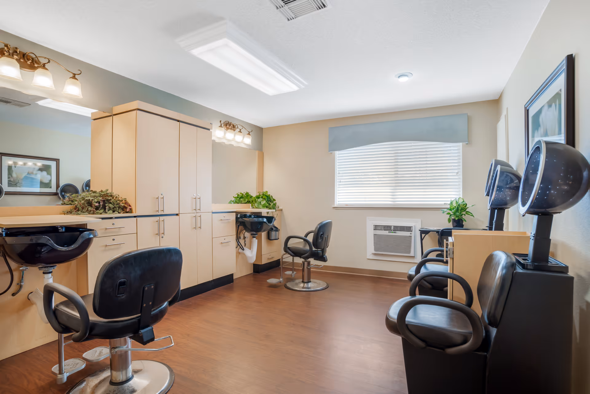 Interior of a hair salon area in a senior living facility with two black salon chairs in front of sinks and mirrors, three black hair dryers on the right side, wooden cabinets, a window with blinds, and plants on the counters.