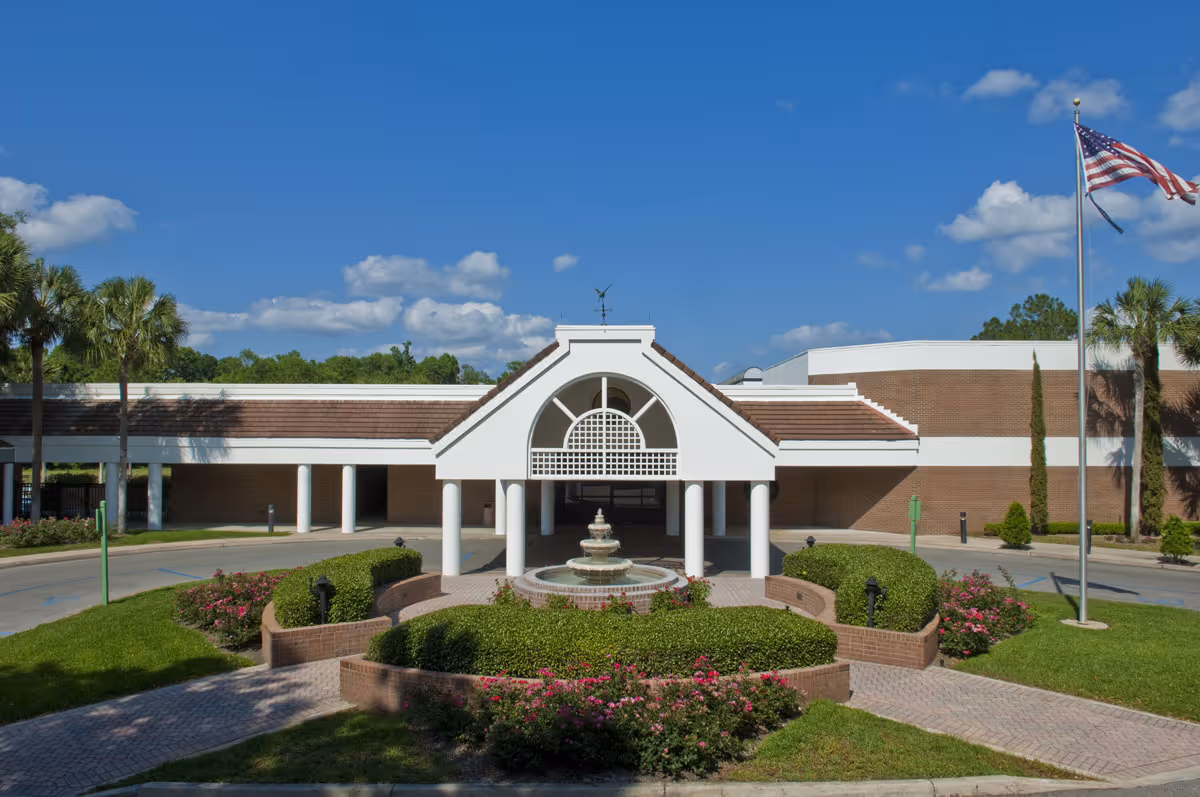 Front exterior view of Village on the Green facility with a covered entrance supported by white columns, a central fountain surrounded by manicured bushes and flowers, palm trees, and an American flag on a flagpole under a blue sky with scattered clouds.