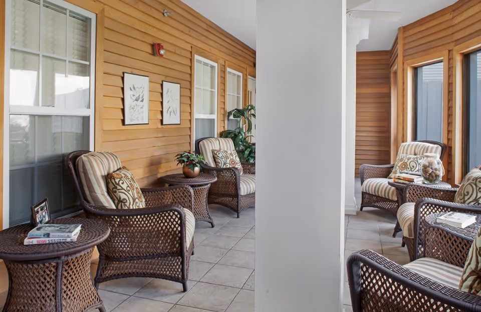 A cozy indoor seating area with wicker chairs featuring striped cushions and patterned pillows, arranged around small wicker tables. The space has large windows, light tiled flooring, and wooden panel walls adorned with framed botanical prints and potted plants.