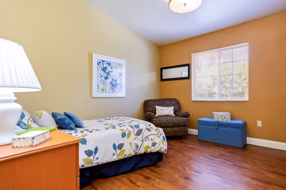 A cozy bedroom with a bed covered in a floral patterned comforter, a wooden nightstand with a white lamp and tissue box, a brown upholstered armchair with a decorative pillow, a blue storage bench, a window with blinds, and framed artwork on the walls.