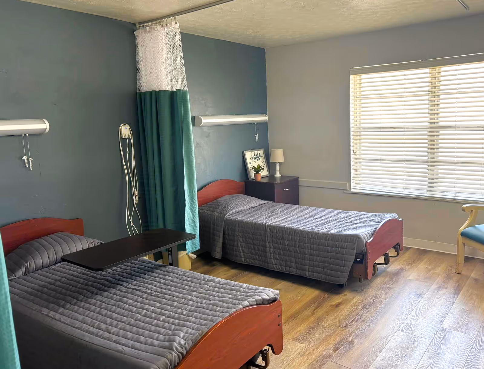 Shared patient bedroom with two single adjustable beds, a privacy curtain, a nightstand and a window with blinds.