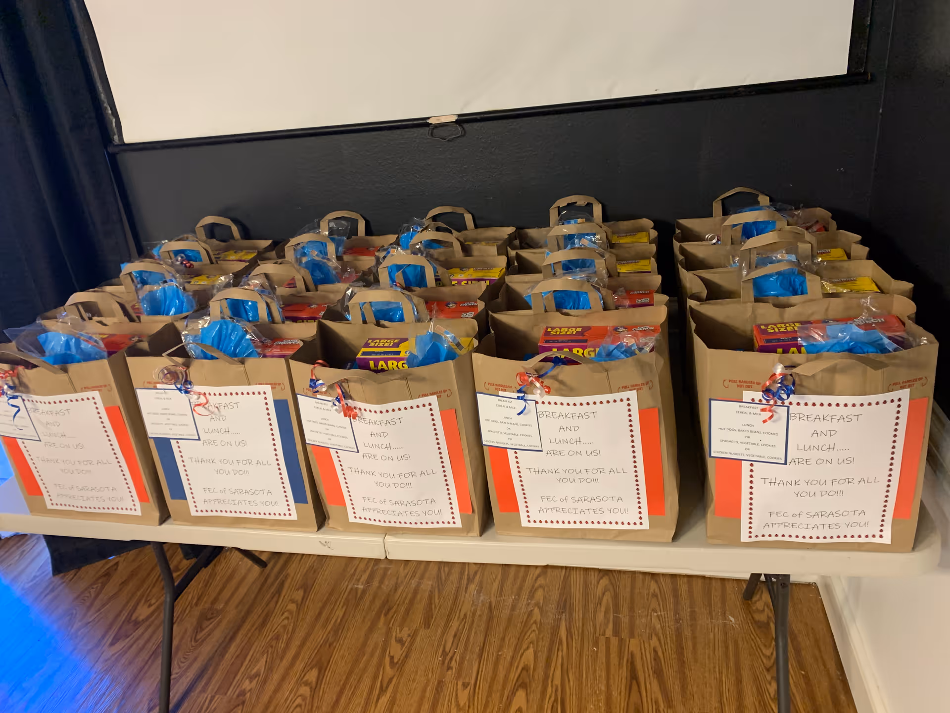 Table lined with rows of paper gift bags filled with packaged items and labeled notes in an indoor room.