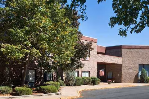 Brick senior living facility exterior with trees, shrubs, and an entrance near an American flag.