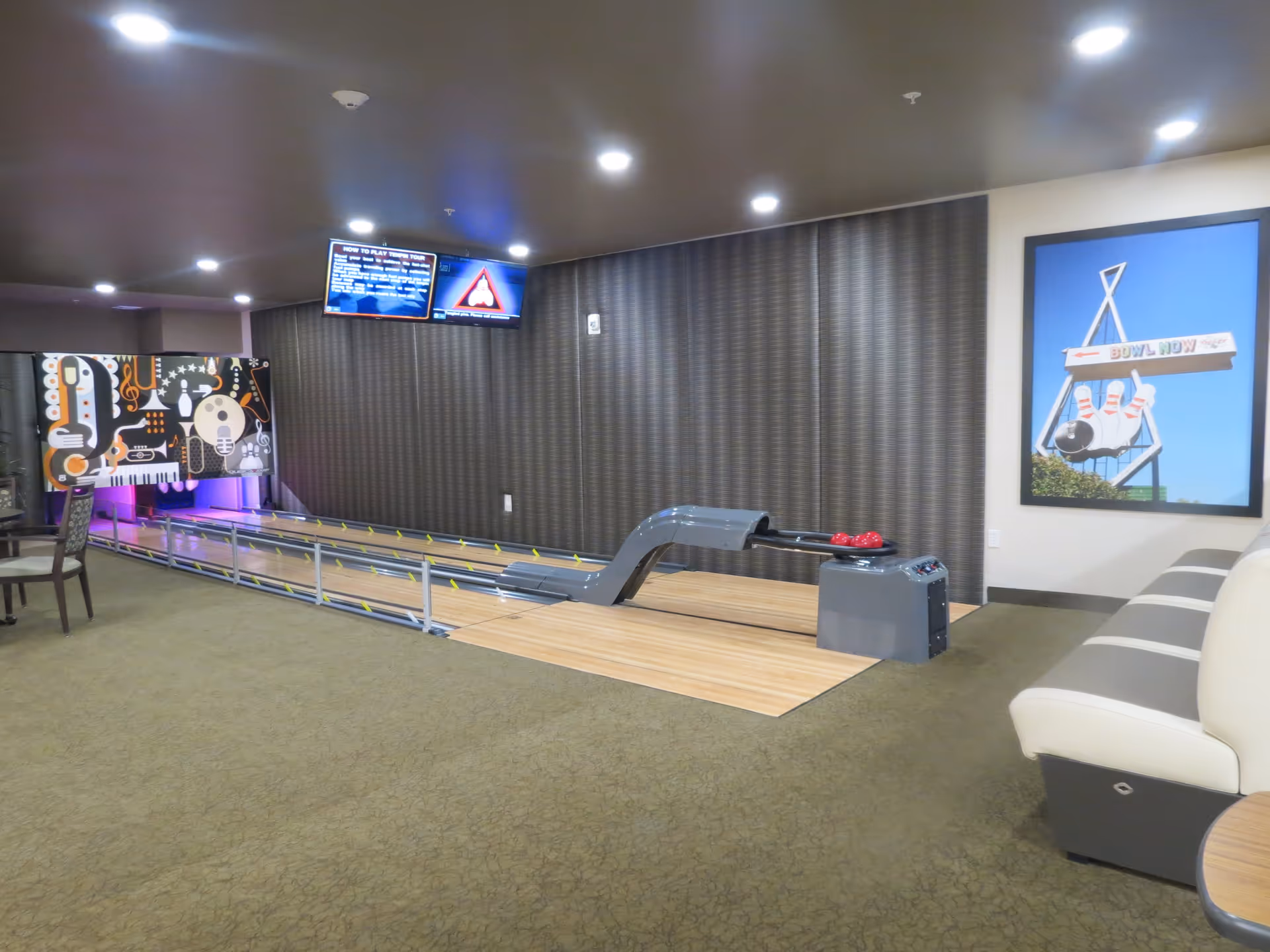 Indoor bowling alley with a single lane, featuring a ball return system with three red bowling balls. The lane has bumpers and a colorful artistic backdrop with musical instruments. There are two screens mounted on the wall above the lane displaying game instructions and scores. To the right, there is a cushioned bench seating area and a framed picture of a bowling sign on the wall.