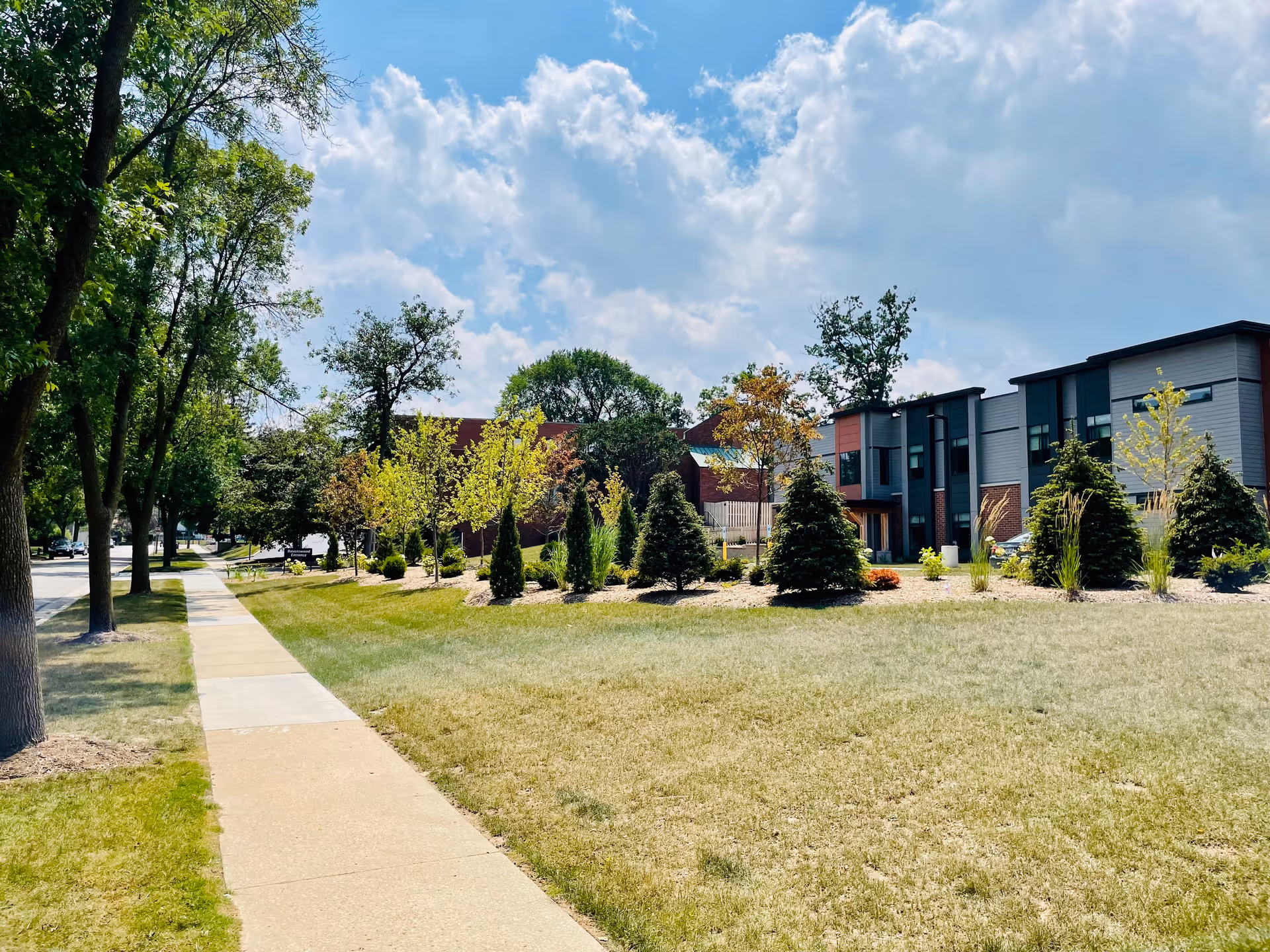 Sidewalk and grassy lawn leading past trees to a modern two-story building with landscaped shrubs under a partly cloudy sky.