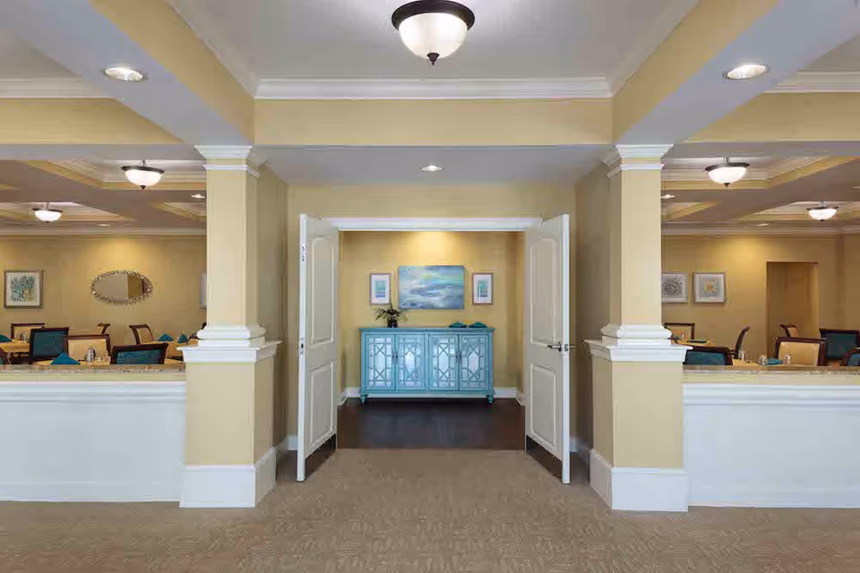 Interior view of a dining room area in a senior living facility with beige walls, white trim, and ceiling lights. The room features multiple tables and chairs with teal napkins, and a blue sideboard with artwork above it is visible through open double doors.