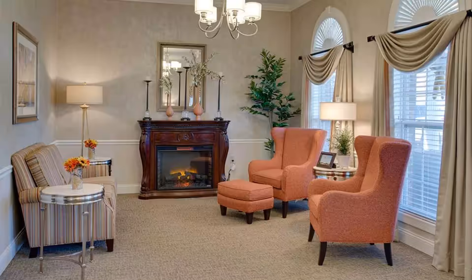 A cozy senior living facility sitting area with two orange armchairs and a matching ottoman, a striped loveseat, a wooden electric fireplace with a mirror above it, two side tables with lamps and flowers, and large windows with draped curtains allowing natural light in.