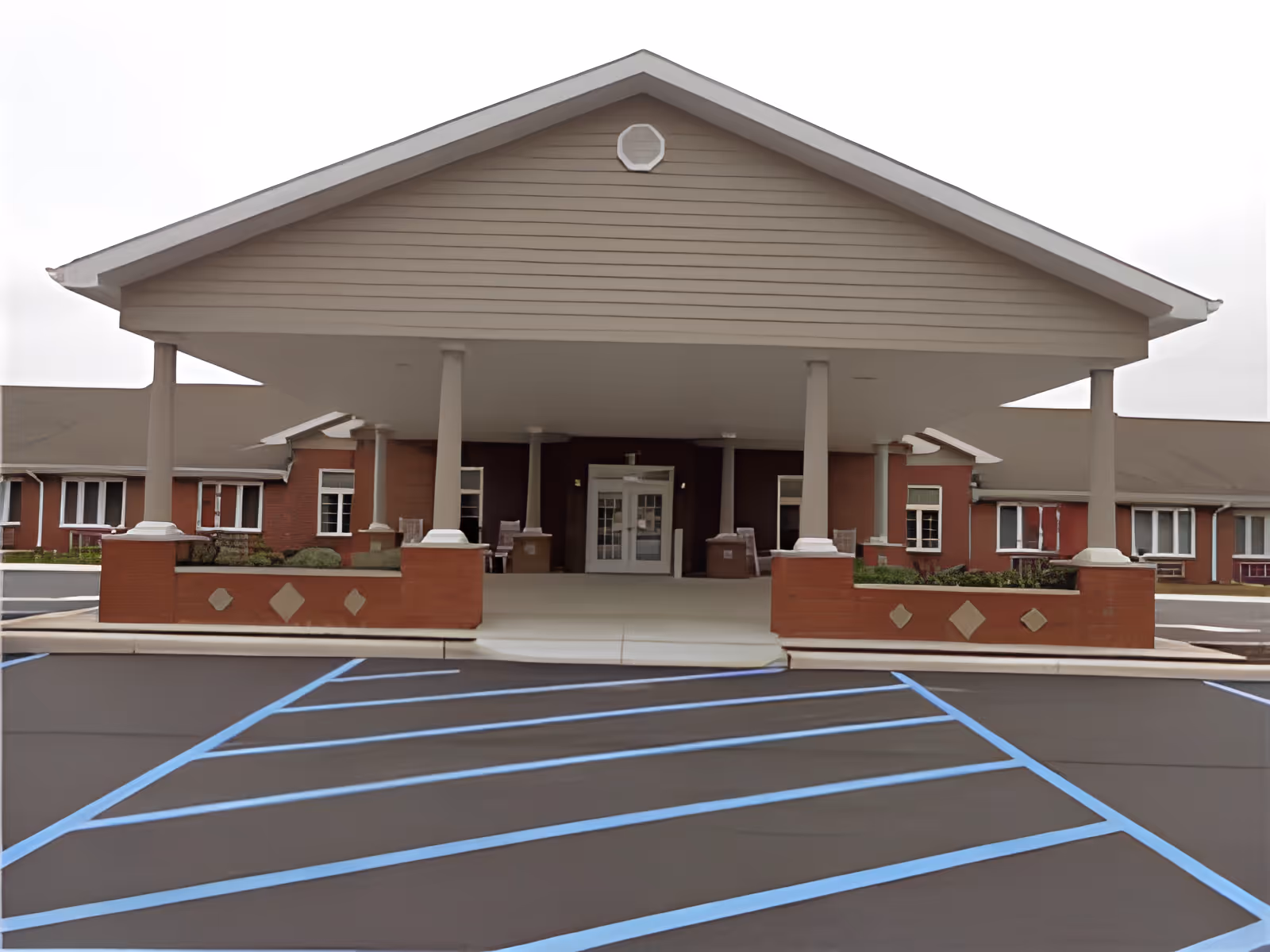 Front exterior view of Grand Valley Gardens Senior Living Community showing a covered entrance with supporting columns, brick planters, and a parking area with blue handicap parking lines.