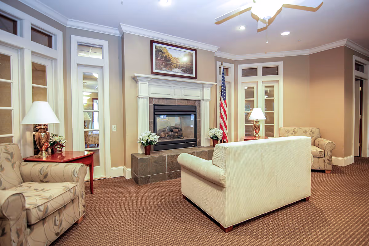 Comfortable common living room with a couch and armchairs arranged around a fireplace, lamps, and an American flag.