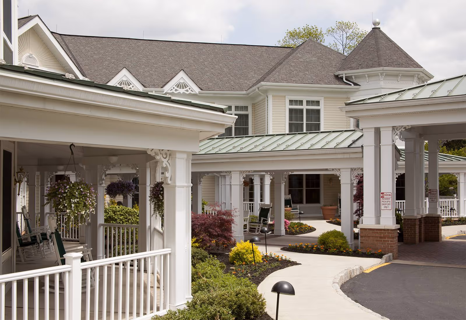 Exterior view of a senior living facility with white wooden columns and railings, a covered porch with hanging flower baskets, rocking chairs, and a landscaped garden with shrubs and flowers along a curved walkway leading to the entrance.