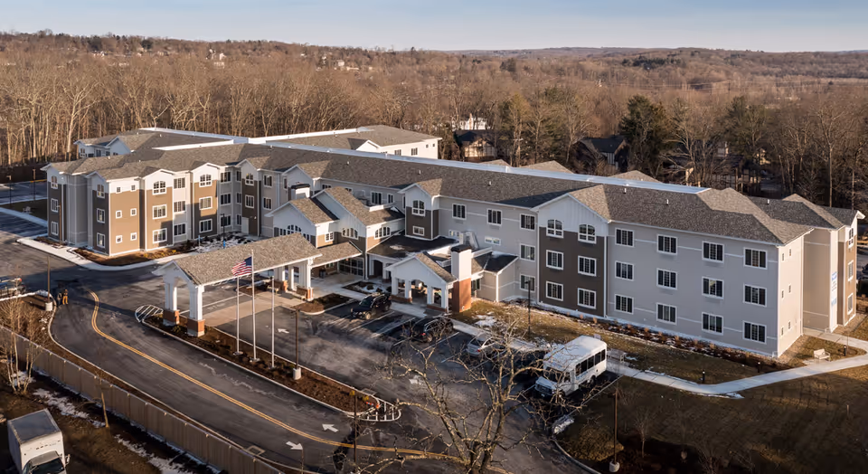 Aerial view of a large, three-story senior living facility building with a covered entrance, multiple windows, and a parking area with several vehicles. The building is surrounded by trees and open land.
