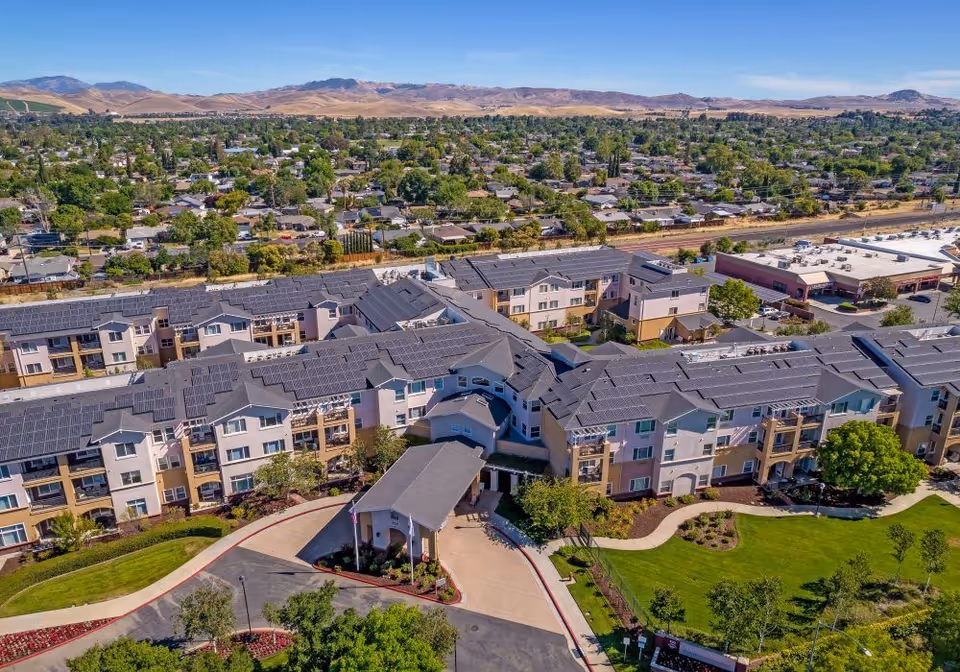 Aerial view of Heritage Estate Senior Apartments, showing a large multi-story residential building with solar panels on the roof, surrounded by landscaped green areas and trees. The building has a covered entrance driveway and is situated in a suburban area with other houses and hills visible in the background under a clear blue sky.