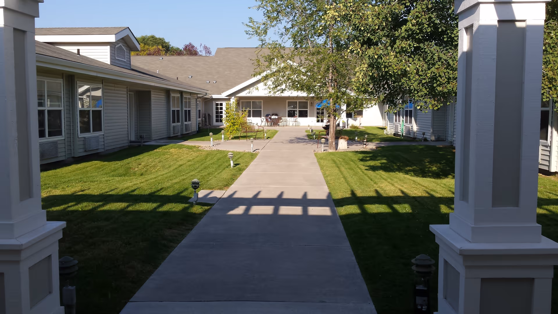View of an outdoor courtyard area in a senior living facility with a concrete walkway flanked by green grass and small garden lights. The courtyard is surrounded by single-story buildings with windows and a few trees providing shade. There are chairs and tables visible in the distance near the building entrance.
