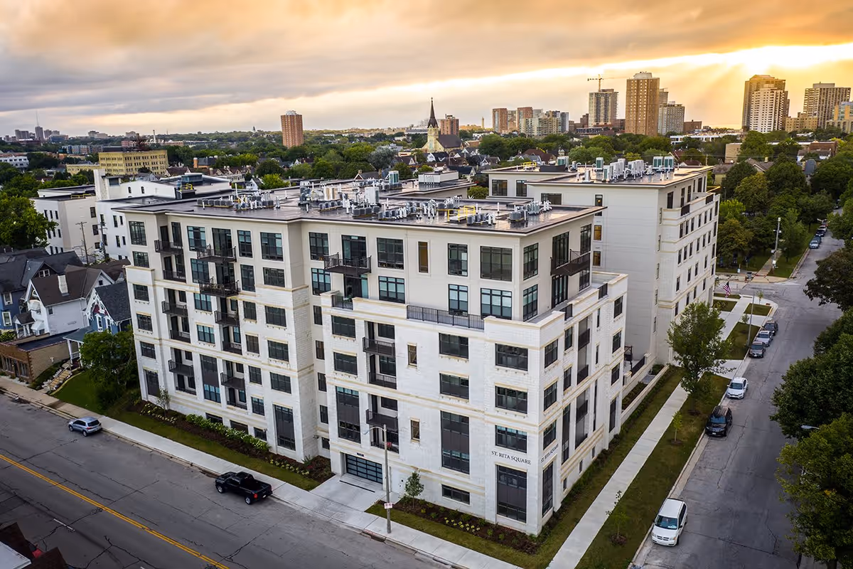 Aerial view of St. Rita Square, a modern multi-story residential building with large windows and balconies, situated in an urban neighborhood with trees and other buildings visible in the background under a cloudy sky at sunset.