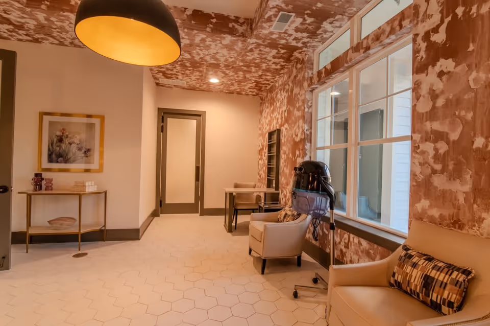Comfortable lounge-style interior with armchairs, a wall-mounted hair dryer, large windows, patterned walls, and a hanging pendant light.